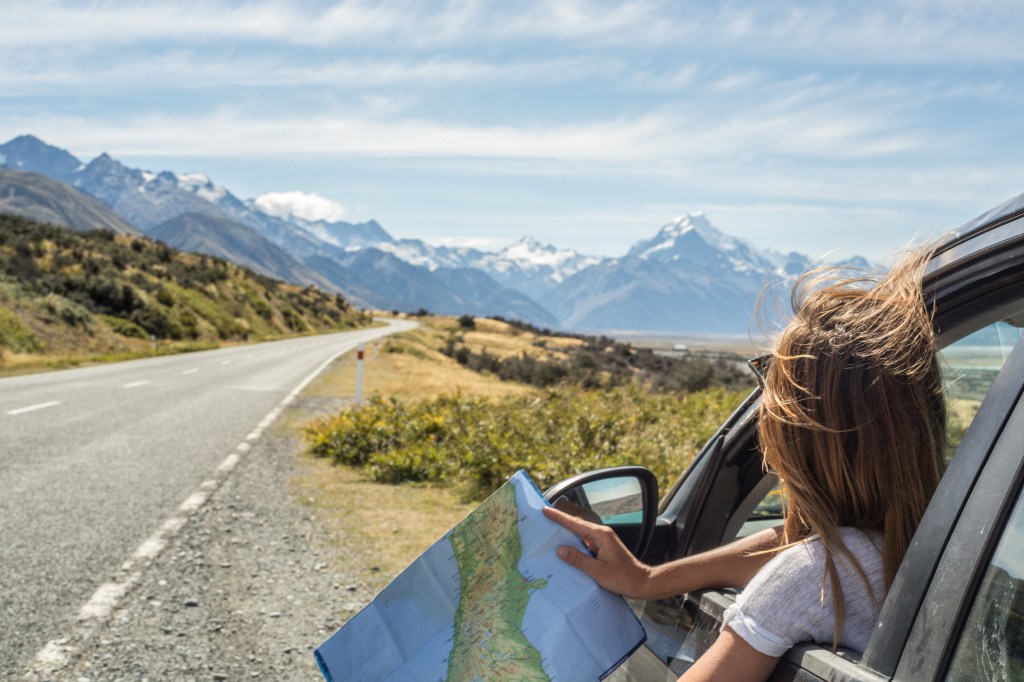 Mulher observa mapa em rodovia com cadeia de montanhas com picos nevados ao fundo