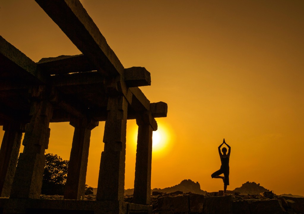 Mulher faz pose de ioga ao lado de uma construção aparentemente abandonada. Ela está contra o pôr-do-sol, de forma que suas feições não estejam visíveis