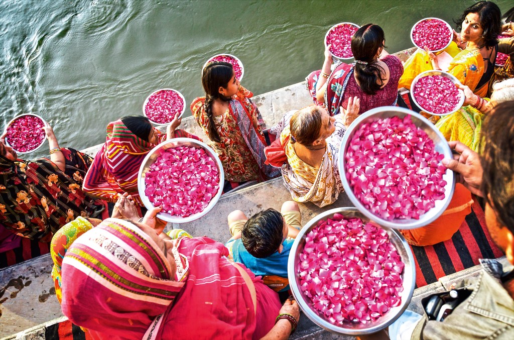 Em degraus de pedra localizados na margem do Ganges, miulheres se reúnem com bacias de pétalas de flores para jogá-las no rio, em oferenda