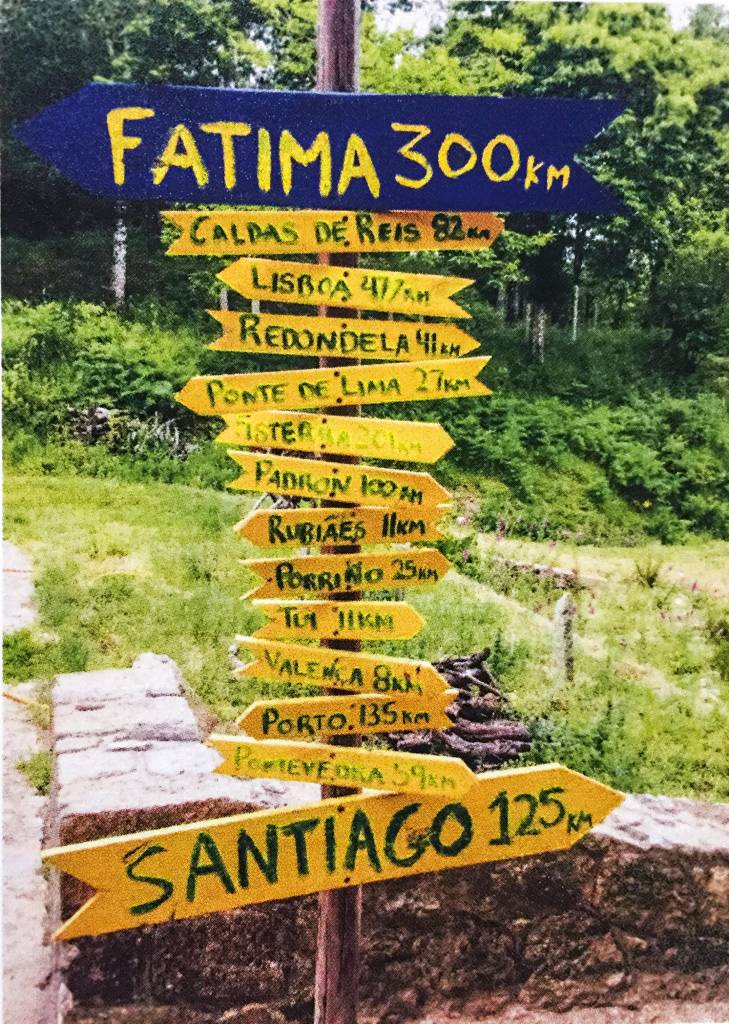 !4 placas em forma de seta, acopladas a um pau vertical, apontam para diversas localizações. Os nomes das cidades estão escritos à mão, entre eles "Santiago 125km", para a direita e "Fatima 300km" para a esquerda.