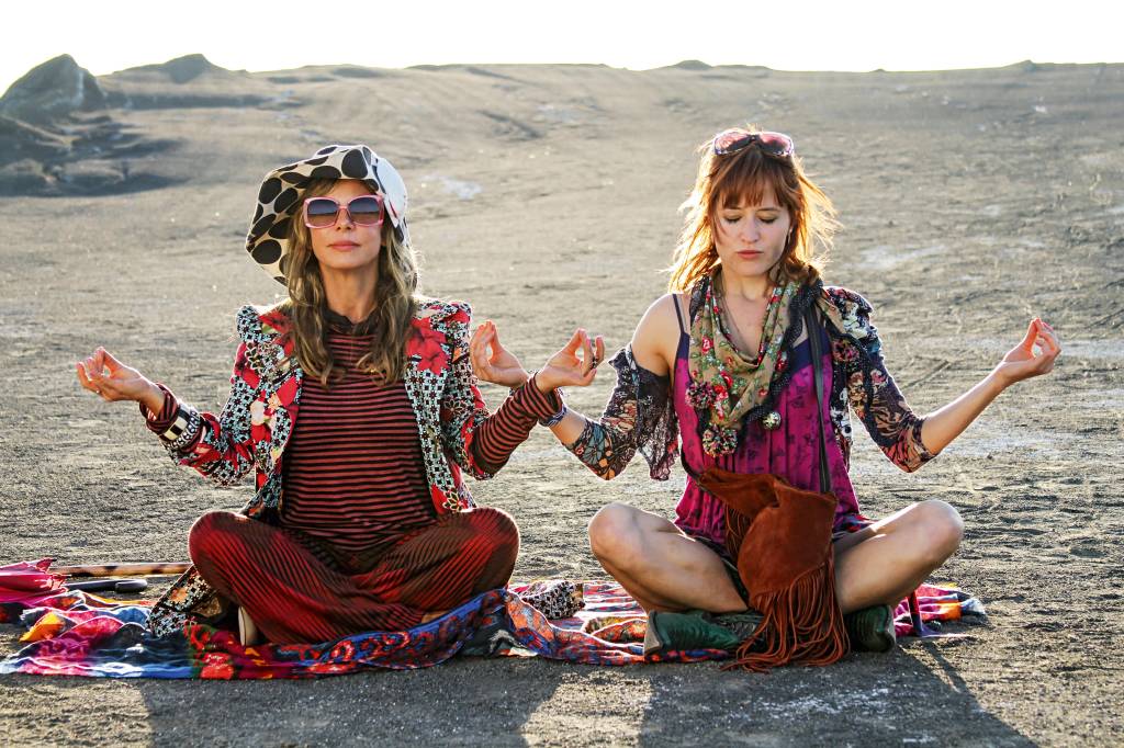 Duas mulheres, vestidas com roupas extravagantes e muitos acessórios, sentam lado a lado, com as mãos levantadas na altura dos ombros em sinal de meditação