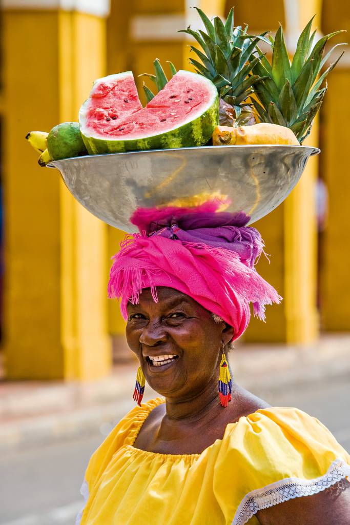 Mulher em trajes de cores fortes carregando uma bacia de frutas em cima da cabeça. É possível distinguir meia melancia, três abacaxis, um abacate, bananas e mais uma fruta.