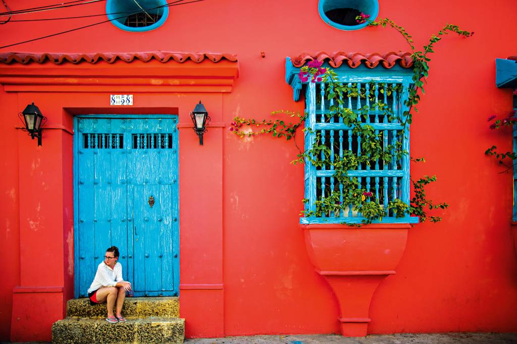 Mulher senta-se na fachada de uma casa típica colombiana, à direita da foto. À esquerda, uma trepadeira surge de dentro da janela