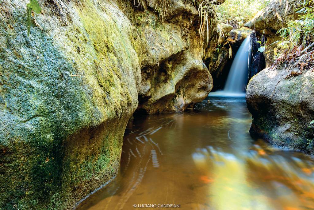 Curso de rio ladeado por pedras altas, com uma pequena cachoeira ao fundo