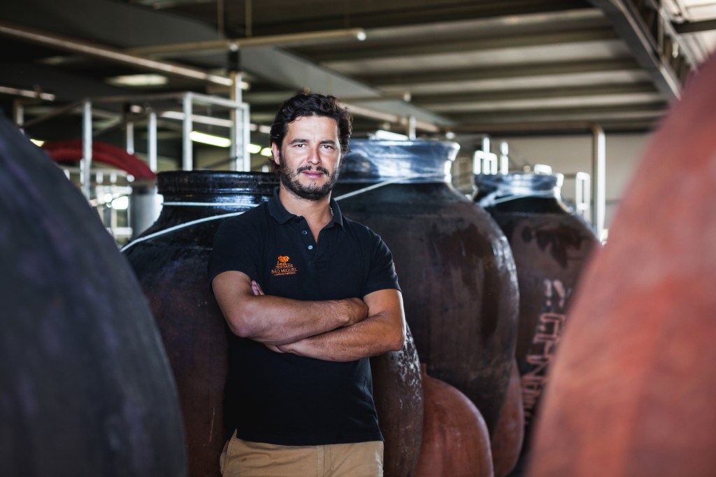 A imagem mostra Alexandre Relvas, um homem branco, com cabelo e barba castanhos. Ele está em pé, em frente de barris de madeira de vinho.