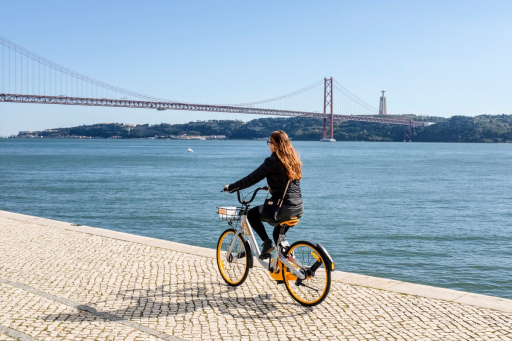 Uma mulher vestida de preto está andando de bicicleta de costas para a câmera. Ao fundo, vemos as águas azuis abundantes do rio Tejo e uma ponte estaiada vermelha.