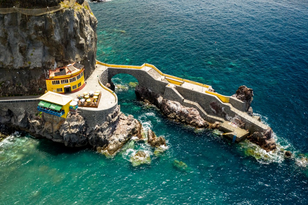 A beira-mar na vila de Ponta do Sol, com uma construção amarela sobre a falésia e pedras no mar, de cor verde esmeralda