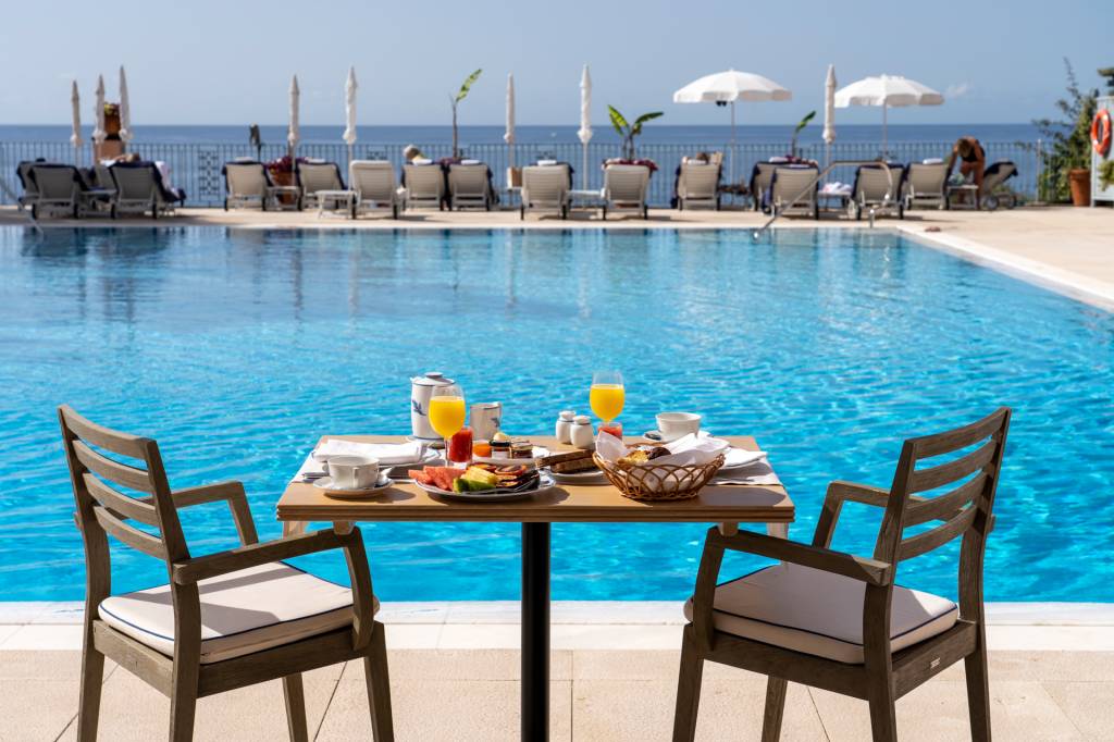 Mesa de café da manhã à beira da piscina do hotel Reid's Palace, na Ilha da Madeira, com duas cadeiras e espreguiçadeiras ao longe