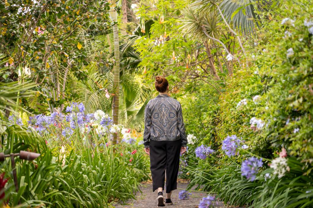 Mulher caminha pelo jardim do hotel Reid's Palace, na Ilha da madeira, entre plantas verdes, flores roxas e árvores