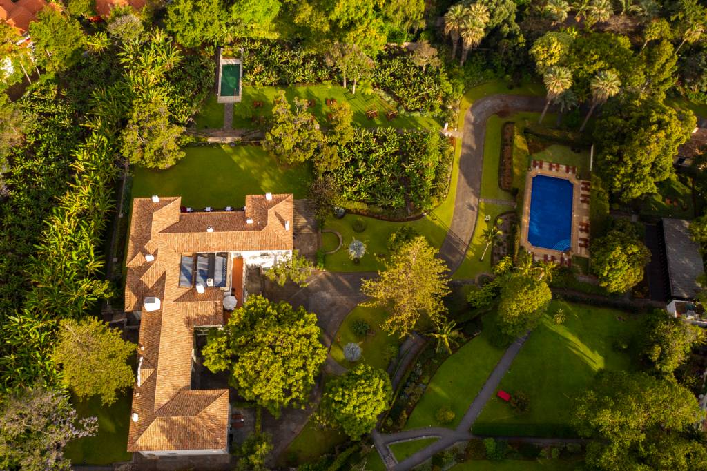 Imagem aérea de uma casa branca cercada de jardins, com uma piscina maior, azul, e uma menor, verde