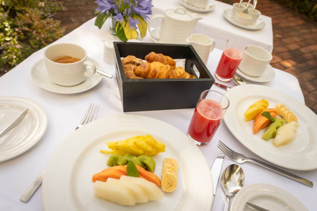 Mesa de café da manhã com prato de frutas, suco vermelho nos copos e cesta de croissants