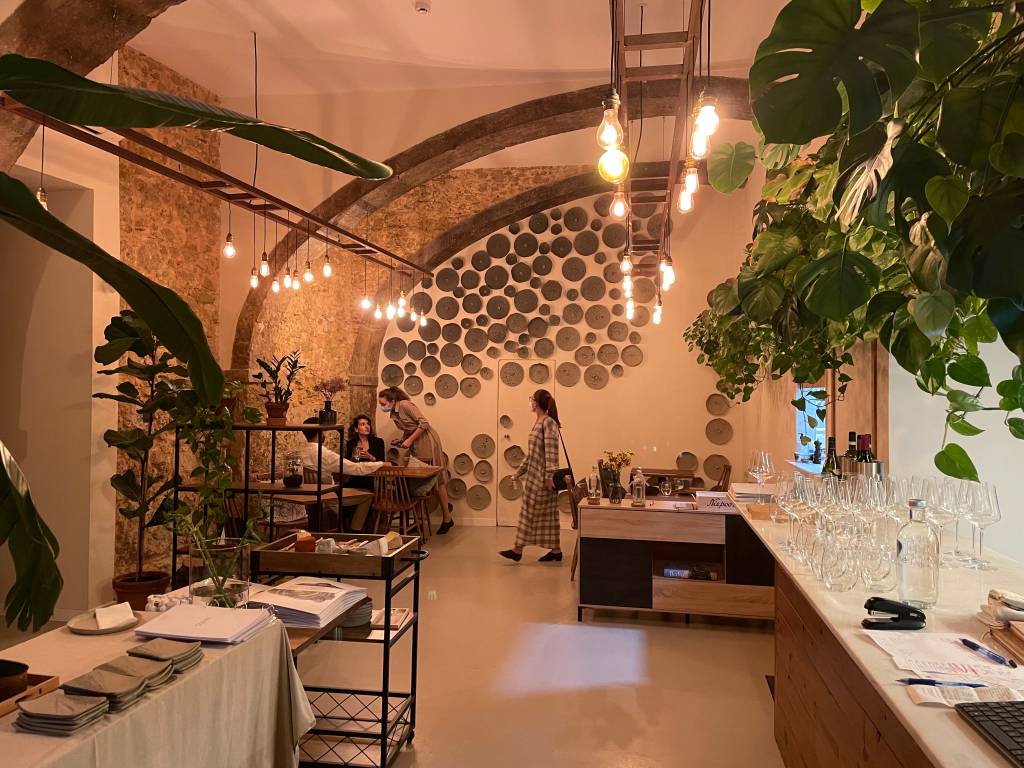 Salão de um restaurante com mesas vazias, plantas à direita, objetos redondos decorando a parede do fundo e uma pessoa a passar na frente.