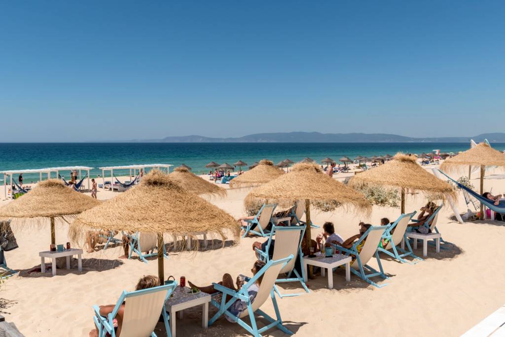 Espreguiçadeiras com guarda-sóis de palha e cadeiras azuis na areia da praia, com o mar azul ao fundo