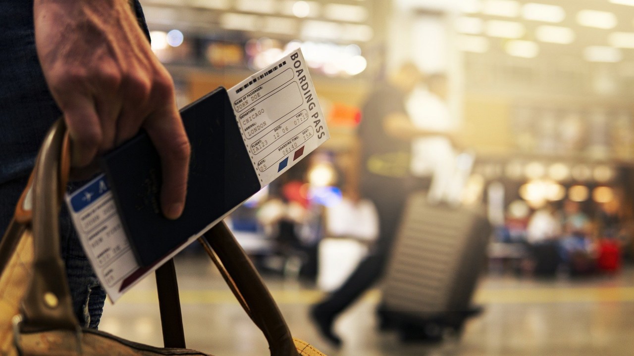 A imagem mostra uma pessoa levando um passaporte, sua passagem e uma mala pelo aeroporto. O fundo está borrado, mas vemos que o aeroporto parece lotado.