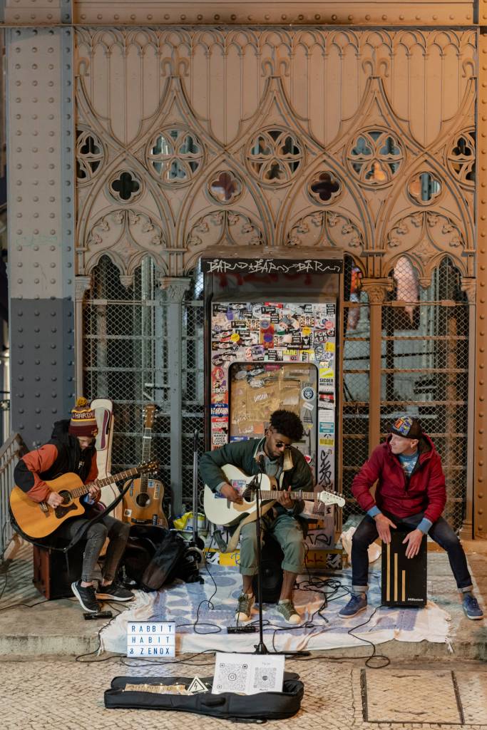 Trio de músicos tocando ao ar livre, encostados numa estrutura de ferro rebuscada