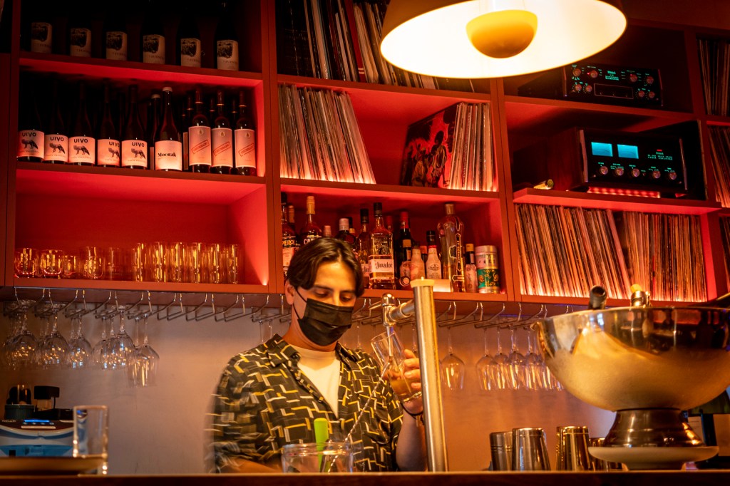 Barman servindo cerveja no balcão de um bar, com uma prateleira atrás recheada de copos, garrafas de bebidas e discos de vinil