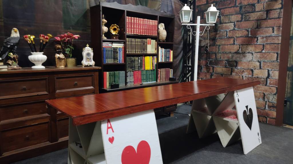 Sala da Escola de Magia Na imagem vemos uma sala onde a mesa foi construída sobre gigantes cartas de baralho e as estantes estão cheias de livros antigos.