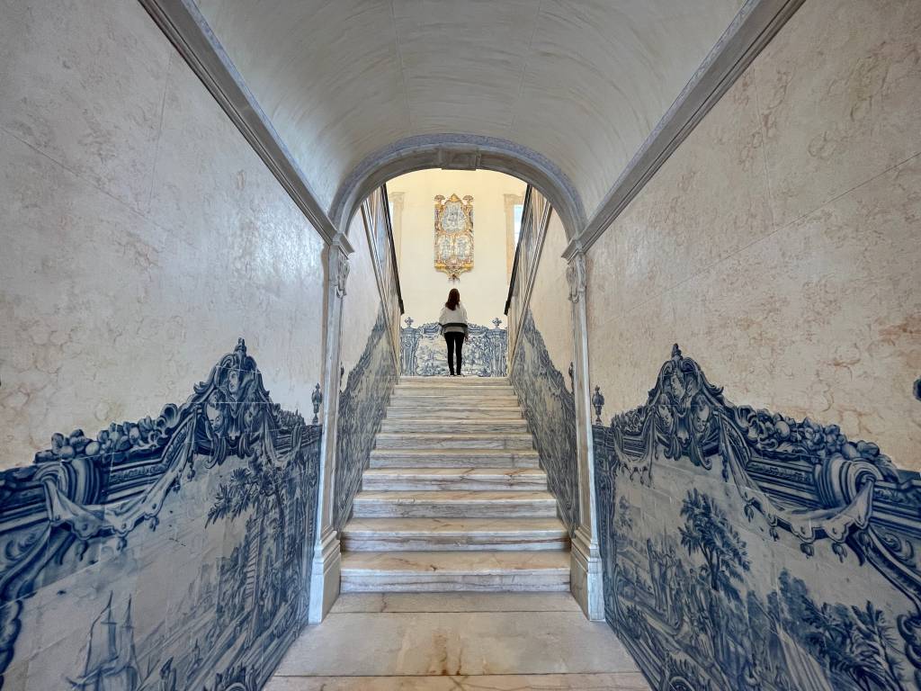 Corredor ladeado de azulejos em azul e branco com um portal e um lance de escadas de mármore, e uma pessoa ao centro