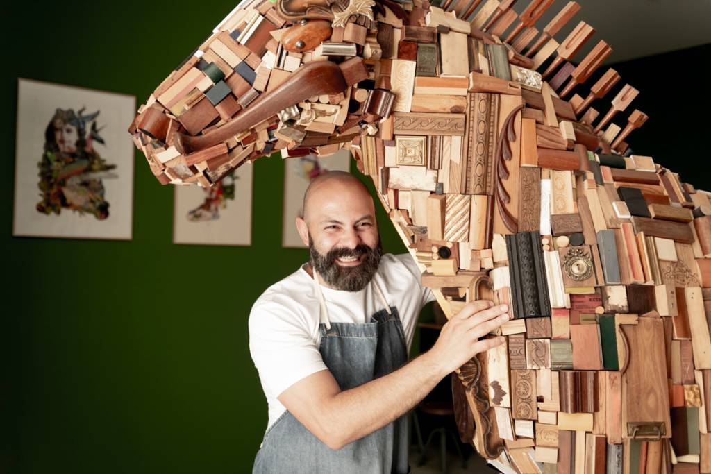 Artista careca, de barba e bigodes, camiseta branca e avental jeans, posa com a sua escultura de cavalo em tamanho real, feita com pedaços de madeira