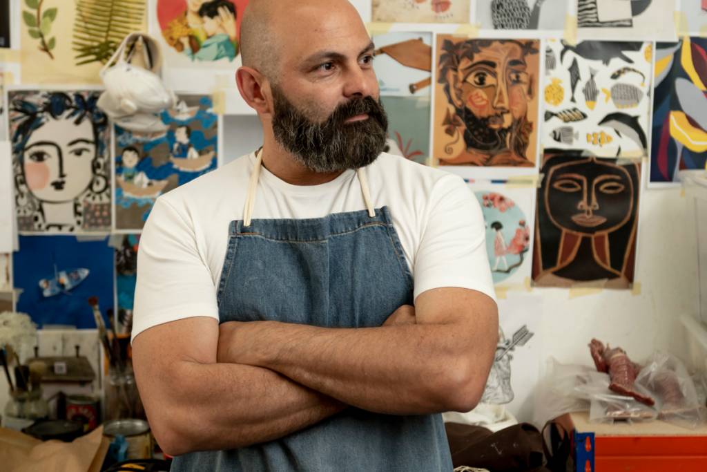 Artista careca, de barba e bigode, posa em seu ateliê, em frente a uma parede cheia de gravuras coloridas