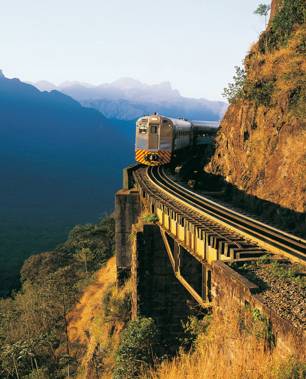 A imagem mostra um trem sobre trilhos curvados. A curva segue o alinhamento de uma montanha ao lado direito, enquanto o lado esquerdo possui um abismo. Ao fundo, a sombra de outras montanhas é azul escuro