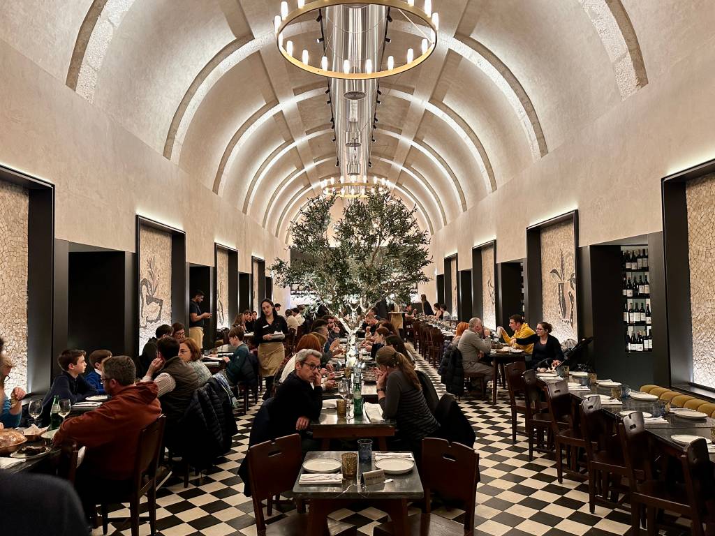 Salão do restaurante com pisco quadriculado em preto e branco sob teto abobadado, onde se vê uma oliveira ao centro