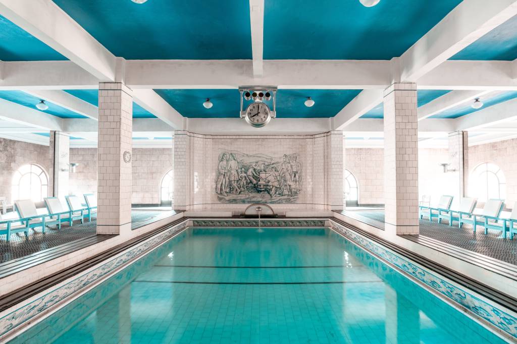 A fotografia colorida mostra uma piscina interna azul. há azulejos brancos ao redor tela e um teto azul.