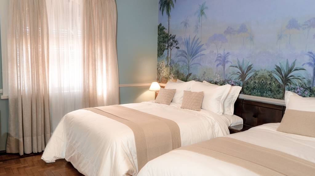 Quarto Superior Deluxe, Grande Hotel Termas de Araxá, Minas Gerais, Brasil A fotografia colorida mostra um quarto com pinturas florais na parede e duas camas de casal com lençóis brancos.