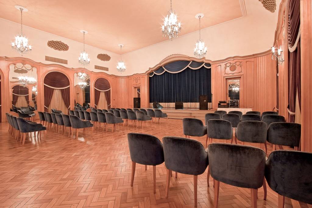 Salão Ouro Preto, Grande Hotel Termas de Araxá, Minas Gerais, Brasil A fotografia colorida mostra um salão com paredes de madeira e um palco com cortina escura. As cadeira aveludadas estão distribuídas como uma plateia