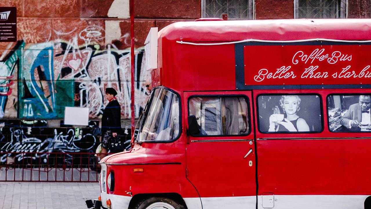 Ônibus antigo vermelho com fotos estampadas em preto e branco estampadas nas janelas. Na lataria, a frase: coffee bus, better than Starbu...