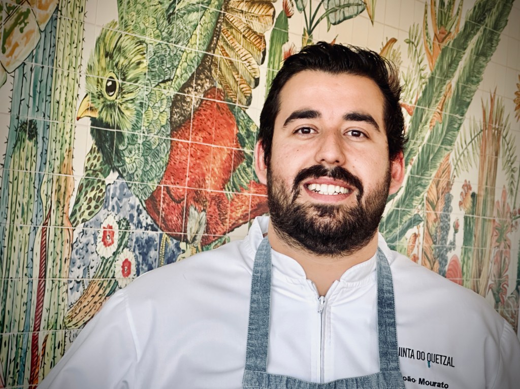 O chef João Mourato, um jovem de barba e cabelos pretos, em frente a um painel de azulejos coloridos