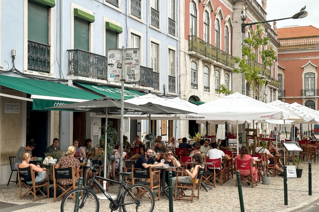 Café com mesinhas na calçada sob ombrelones, de frente para as barracas da Feira da Ladra