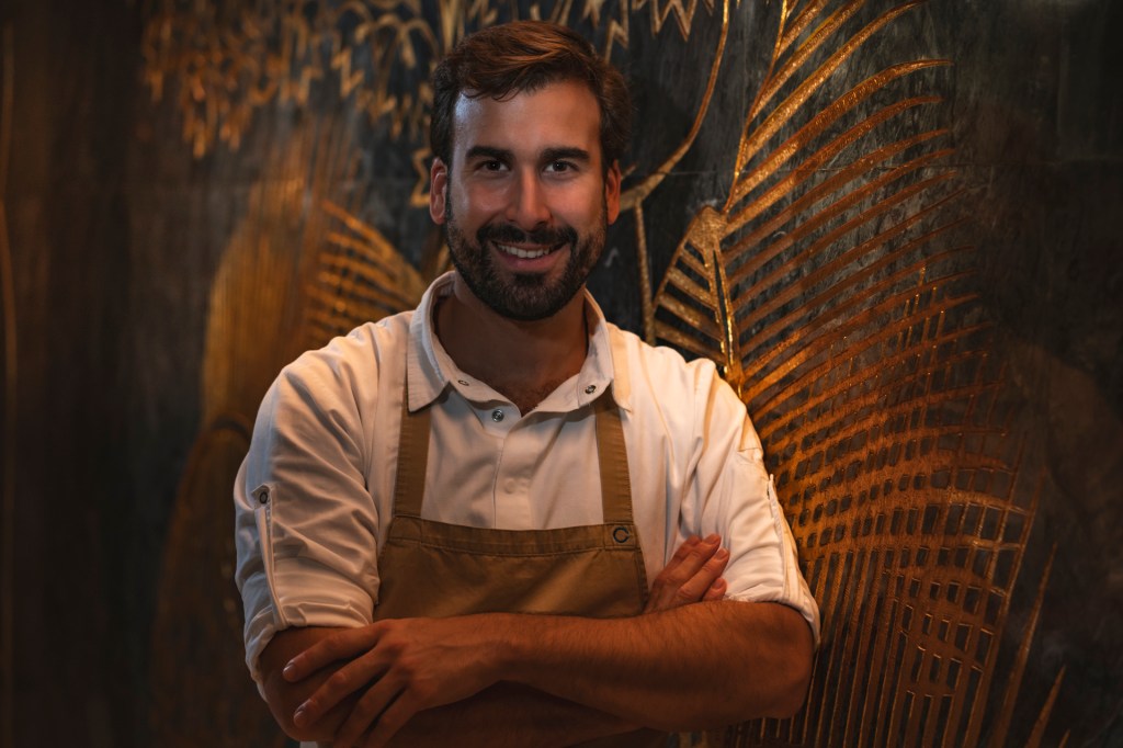 Fotografia colorida. Ocupando o centro da imagem, o jovem chef Pedro Pena Bastos, vestido de camisa branca e avental marrom