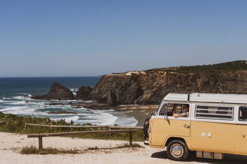 Foto colorida mostra uma praia selvagem com uma kombi antiga em primeiro plano