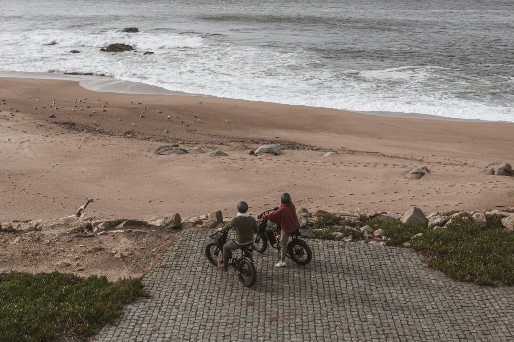 Fotografia colorida de drone mostra um casal de bicicleta parado contemplando o mar