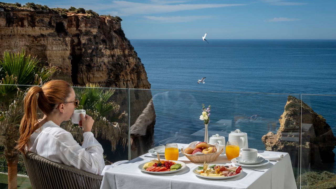 Mulher toma uma xicara de café sentada numa mesa onde o café da manhã está servido, numa varanda de vidro com vista da falésia e do mar