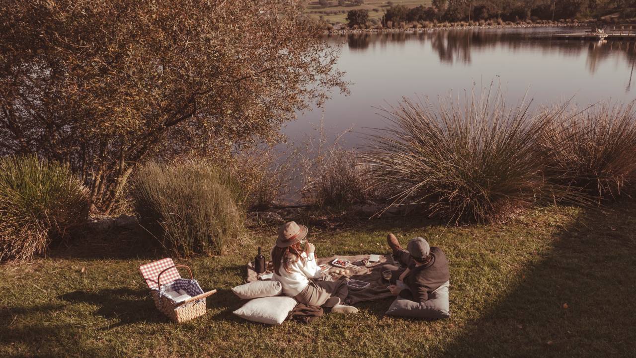 A imagem mostra um casal fazendo piquenique na beira do lago, com uma cesta recheada de guloseimas da região e uma garrafa de vinho