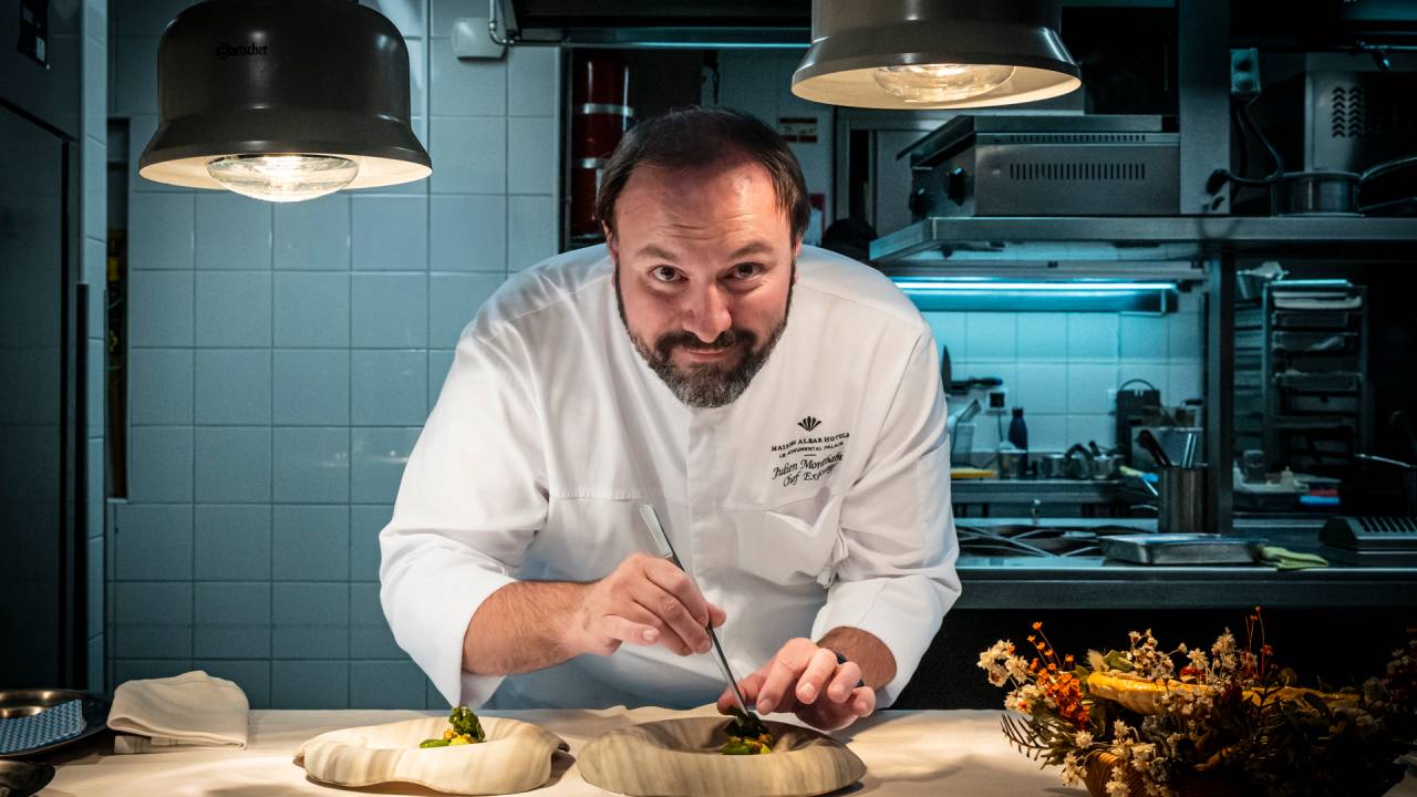 Em uma bancada na sua cozinha, o chef, vestido com o seu jaleco, finaliza um prato com uma pinça